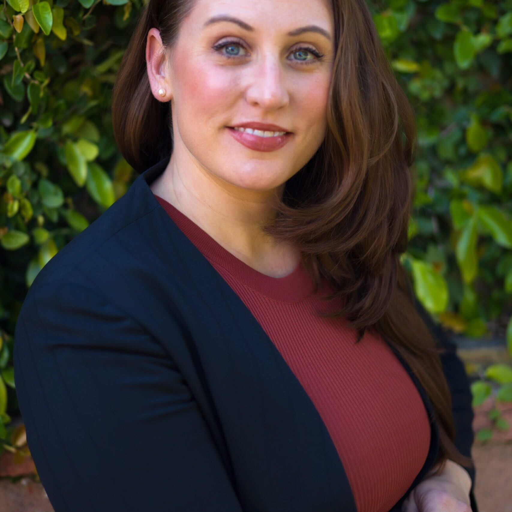 Smiling woman with long brown hair wearing a black blazer and red top outdoors.